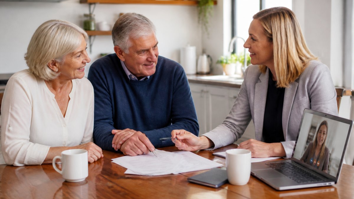 A mature couple in their late 50s sitting at a kitchen table with a financial adviser, reviewing documents and smiling, with a laptop open showing a video call